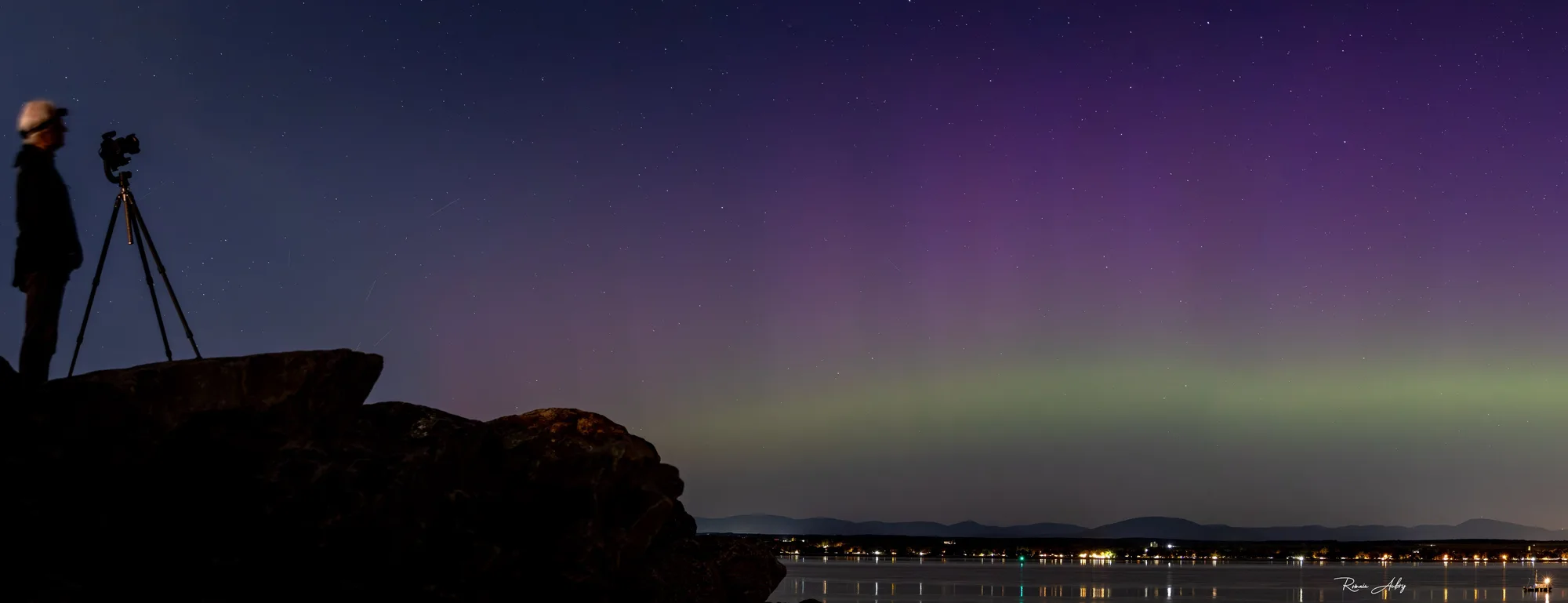 An aurora chaser standing beside a camera tripod on coastal rocks, watching a green auroral band and purple pillars over distant town lights.