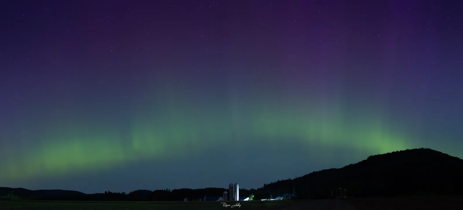 A green auroral arc low over a farm with silos, beneath a purple sky — a typical mid-latitude display seen from Quebec.